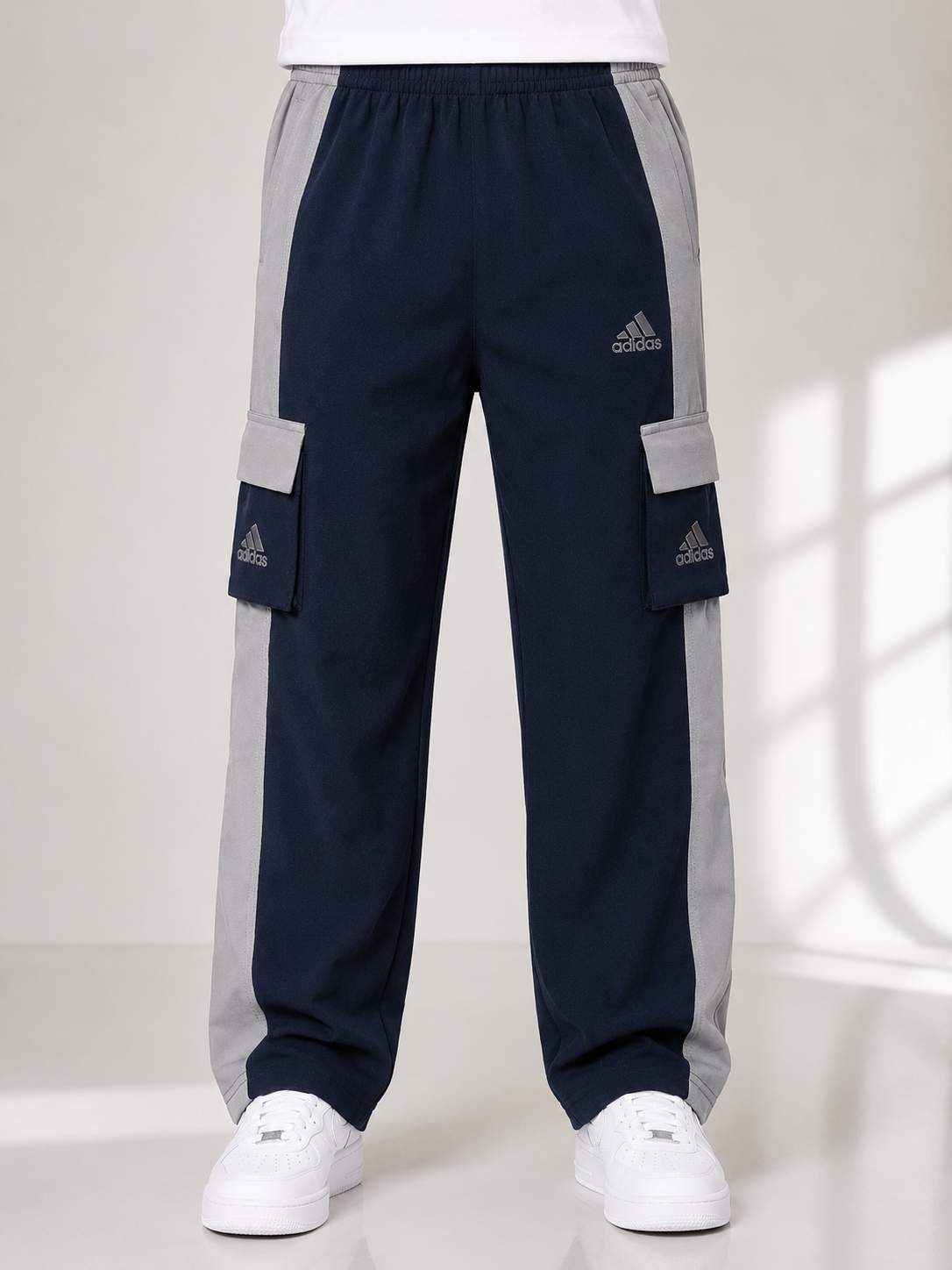 A person wearing navy blue Adidas cargo jogger pants with light gray side panels and white sneakers, shown from the waist down against a light studio background.
